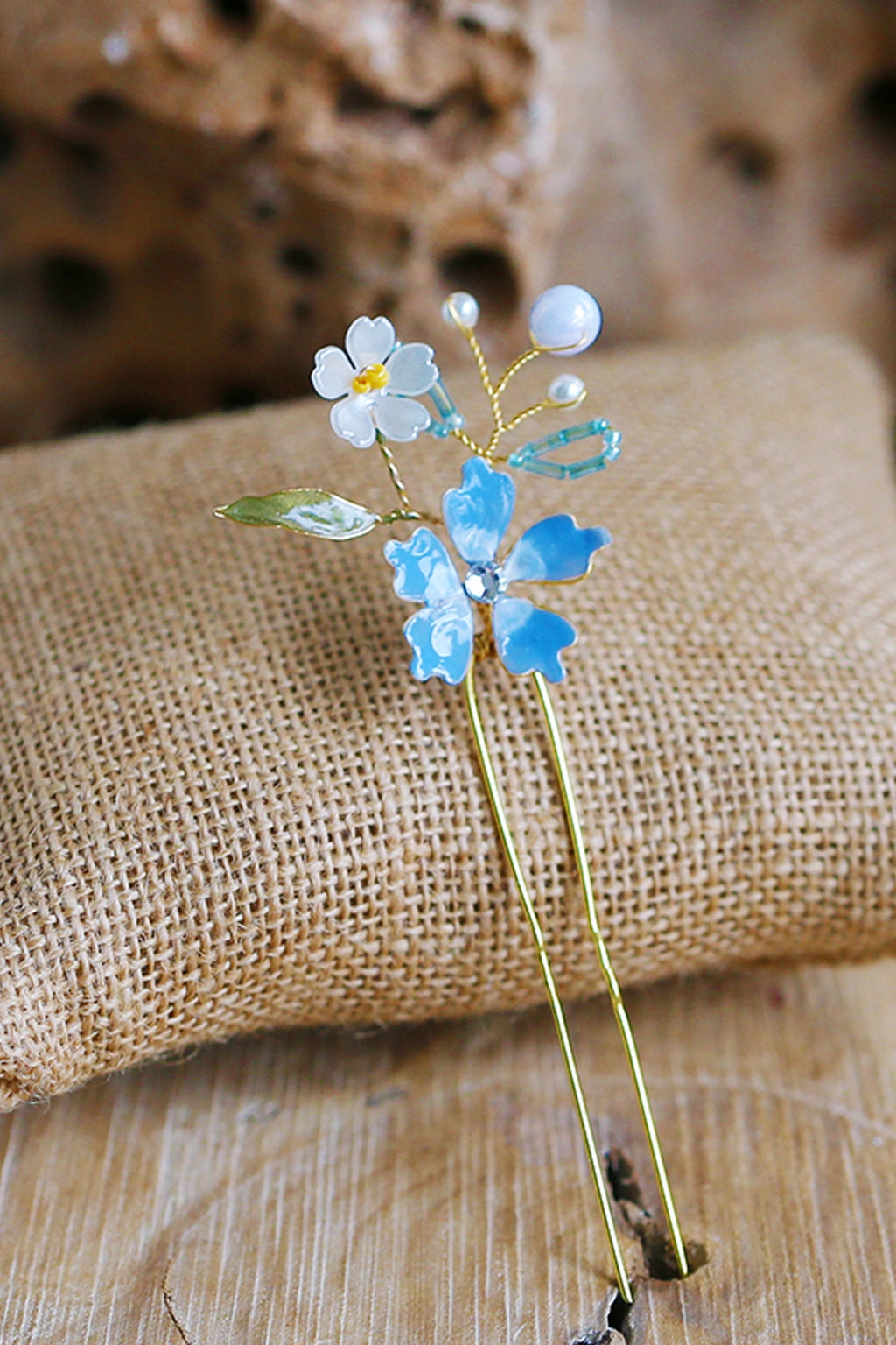 Handmade blue resin floral bridal hair pin with pearls and gold wire detailing displayed on rustic fabric background.
