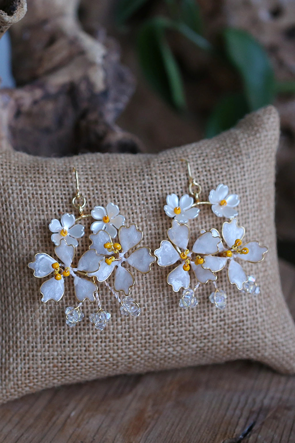 Handmade white resin floral earrings with gold-plated wire and crystal danglers displayed on burlap cushion
