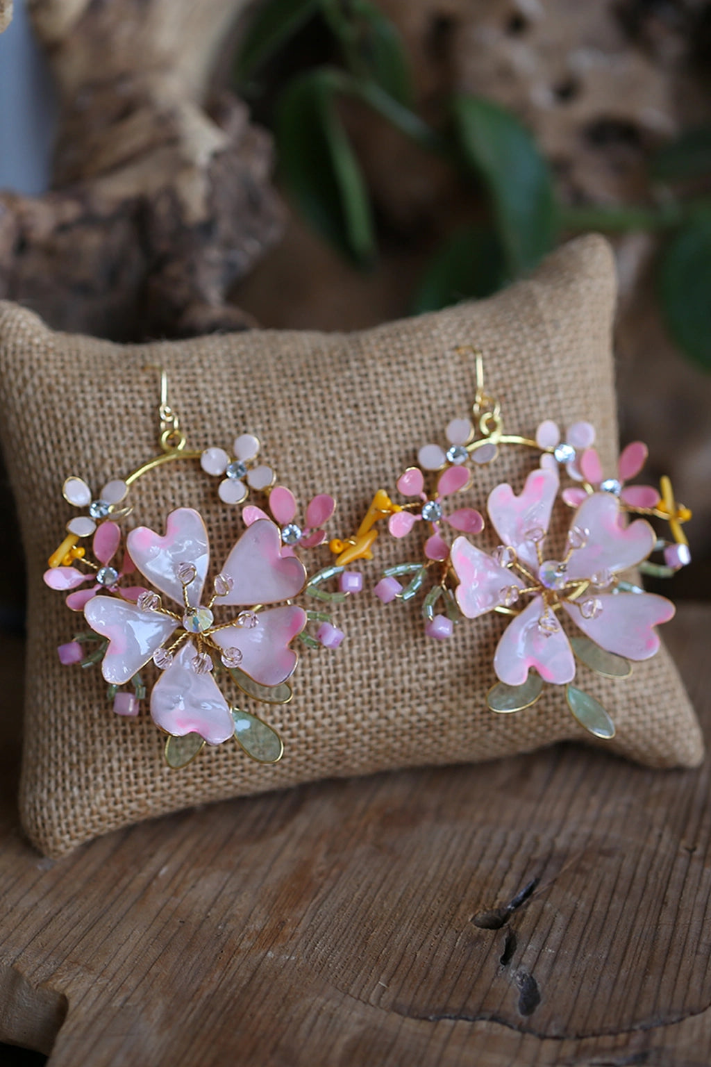 Handmade pink floral resin bridal earrings with pearls, crystals, and gold metal detailing displayed on jute cushion.