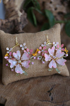 Handmade pink floral resin bridal earrings with pearls, crystals, and gold metal detailing displayed on jute cushion.