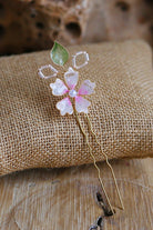 Handmade blush pink resin floral bridal hair pin with pearl centre and gold wire detailing displayed on rustic fabric background.